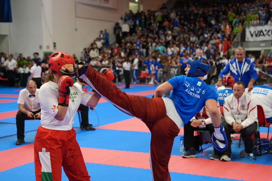 Kickboxerin mit blauer Kleidung tritt gegen Gegnerin mit rotem Helm in einer Sporthalle in Österreich.