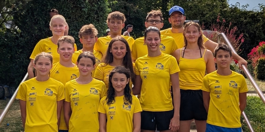 Jugendliche und Erwachsene in gelben Trikots posieren für ein Gruppenfoto vor blühenden Pflanzen.