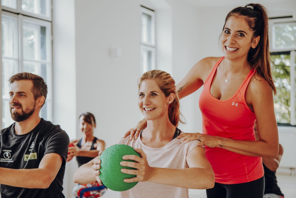 Trainerin steht hinter Frau mit Gymnastikball